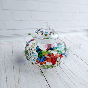 Puede incluir: Una tetera de vidrio transparente con tapa y asa. La tetera est&aacute; decorada con colibr&iacute;es, flores y enredaderas pintados a mano. El dise&ntilde;o incluye tonos de azul, verde, rojo y naranja. La tetera se encuentra sobre una superficie de madera clara.