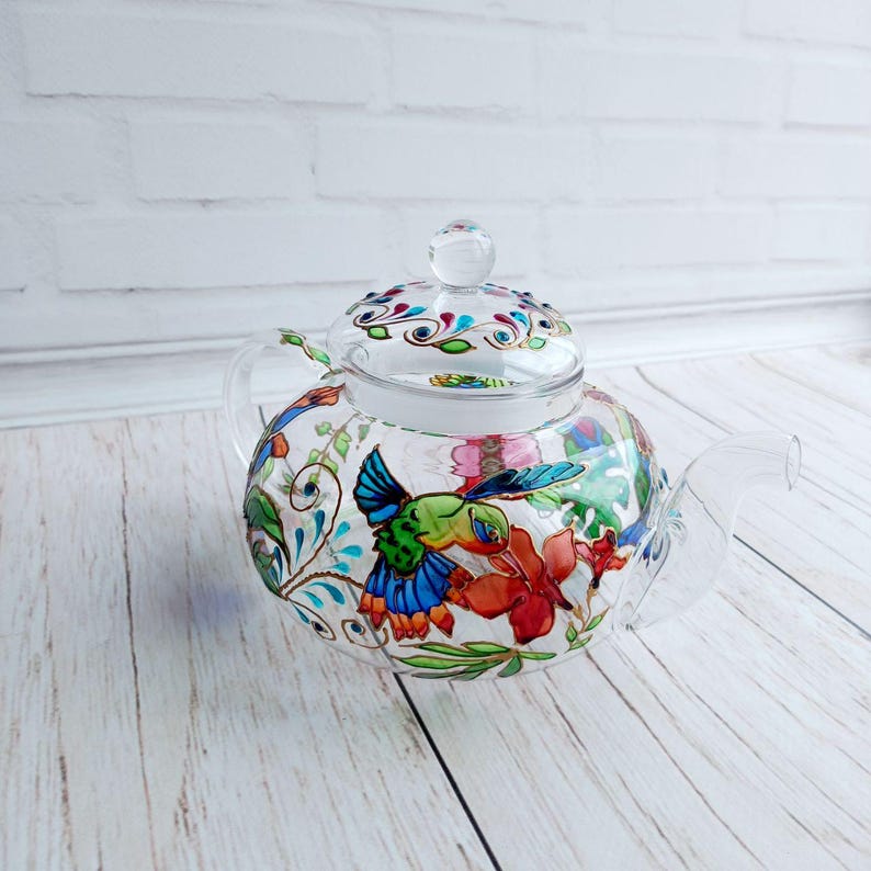 Puede incluir: Una tetera de vidrio transparente con tapa y asa. La tetera est&aacute; decorada con colibr&iacute;es, flores y enredaderas pintados a mano. El dise&ntilde;o incluye tonos de azul, verde, rojo y naranja. La tetera se encuentra sobre una superficie de madera clara.