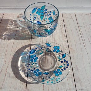 May include: A clear glass teacup and saucer set. The cup and saucer are decorated with a hand-painted floral design in shades of turquoise and blue. The floral pattern includes stylized flowers and leaves, with silver accents.
