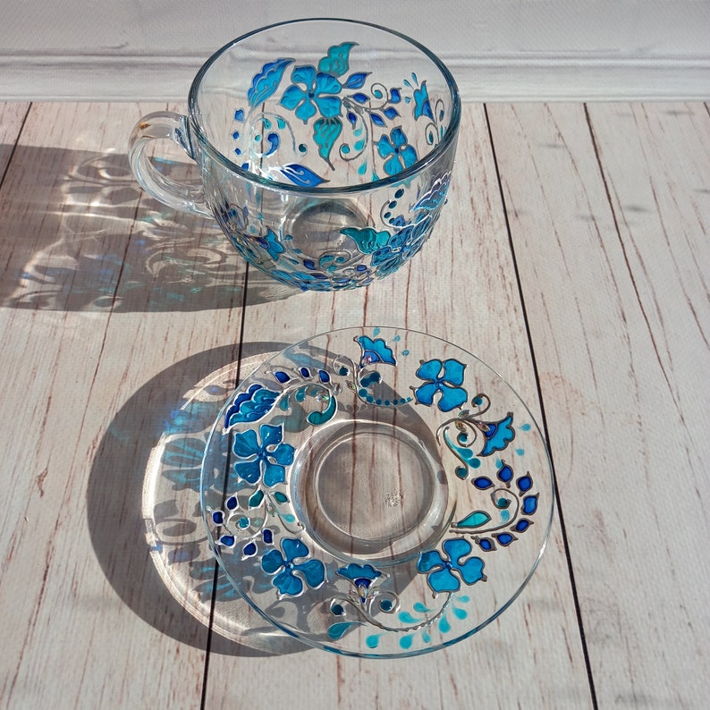 May include: A clear glass teacup and saucer set. The cup and saucer are decorated with a hand-painted floral design in shades of turquoise and blue. The floral pattern includes stylized flowers and leaves, with silver accents.