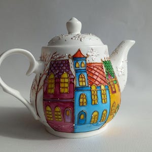 Op de afbeelding: Witte keramische theepot versierd met een kleurrijk dorpsgezicht. De huizen hebben rode, blauwe en gele daken en muren. Het handvat en de tuit zijn wit en het deksel heeft een kleine knop. Een charmant decoratief item.