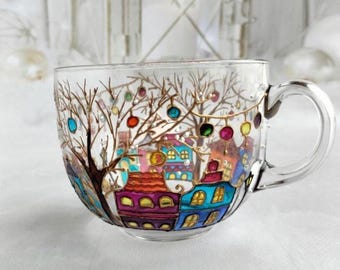 May include: A clear glass mug featuring a hand-painted winter scene with colorful houses in shades of purple, blue, and red, bare trees with gold branches, and hanging multi-colored circular ornaments against a snowy backdrop.