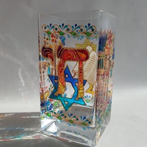 Puede incluir: Un portavelas de vidrio transparente con símbolos judíos pintados a mano. El diseño incluye una estrella de David azul y la palabra hebrea "Chai" en naranja y azul. Patrones florales y detalles dorados decoran los bordes.