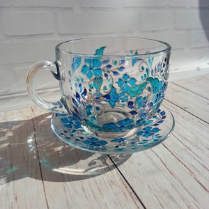 May include: A clear glass teacup and saucer set. The cup and saucer are decorated with a hand-painted floral design in shades of turquoise and blue. The cup has a handle and sits on a matching saucer, creating an elegant tea set.