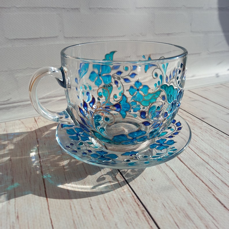 May include: A clear glass teacup and saucer set. The cup and saucer are decorated with a hand-painted floral design in shades of turquoise and blue. The cup has a handle and sits on a matching saucer, creating an elegant tea set.