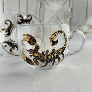 May include: A clear glass teacup and matching saucer set featuring a hand-painted scorpion design. The scorpion is rendered in black and metallic gold with intricate swirling patterns. The text Scorpio is partially visible on the glass surface.