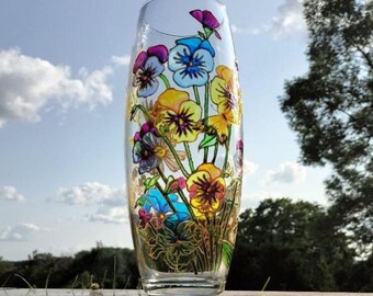 May include: A clear glass vase with a hand-painted floral design featuring colorful pansies in shades of purple, blue, and yellow. The vase is cylindrical and stands on a reflective surface, with a backdrop of a blue sky and greenery.