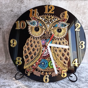 May include: A decorative black owl clock with gold numerals and hands. The owl design features intricate gold, blue, and red detailing. The clock is displayed on a black metal stand, adding a touch of elegance to any room.