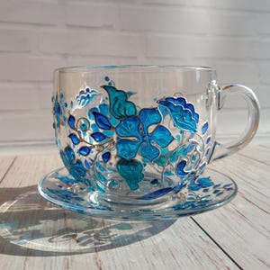 May include: A clear glass teacup and saucer set, hand-painted with vibrant blue floral designs. The cup features a large handle and sits on a matching saucer, creating an elegant and artistic tea set.