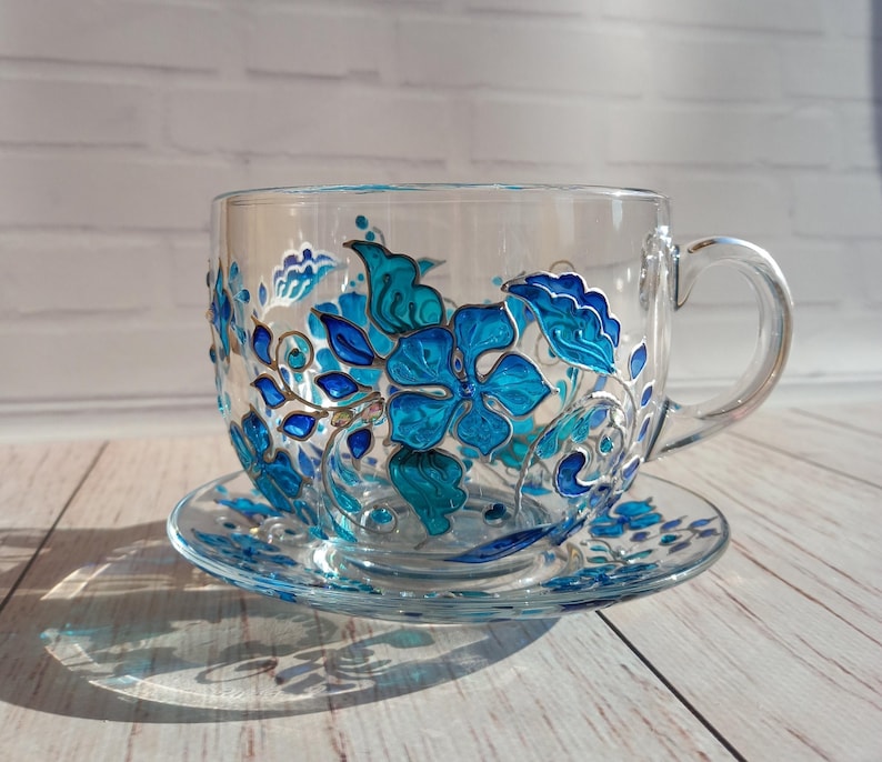 May include: A clear glass teacup and saucer set, hand-painted with vibrant blue floral designs. The cup features a large handle and sits on a matching saucer, creating an elegant and artistic tea set.