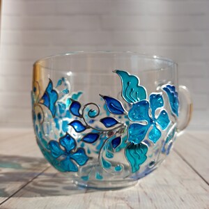May include: A clear glass teacup with a handle, adorned with hand-painted blue and teal floral designs. The flowers and leaves are outlined in silver, creating a decorative and elegant look. The cup is set against a light-colored wooden surface.