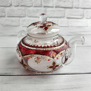 Può includere: Teiera in vetro trasparente con coperchio e manico. La teiera è decorata con disegni floreali rossi, oro e bianchi. Il coperchio ha un pomello in vetro trasparente. La teiera è appoggiata su una superficie di legno bianco.