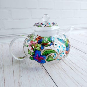 Puede incluir: Una tetera de vidrio transparente con tapa, asa y boquilla. La tetera est&aacute; decorada con coloridos dise&ntilde;os florales y de colibr&iacute;es en azul, rosa, verde y dorado. La tetera se encuentra sobre una superficie de madera clara.