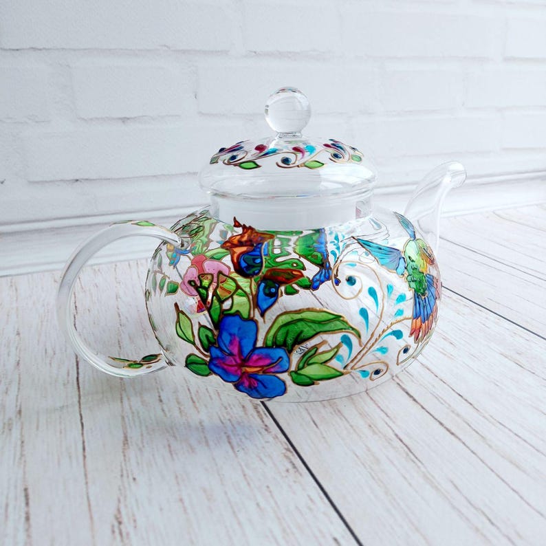 Puede incluir: Una tetera de vidrio transparente con tapa, asa y boquilla. La tetera est&aacute; decorada con coloridos dise&ntilde;os florales y de colibr&iacute;es en azul, rosa, verde y dorado. La tetera se encuentra sobre una superficie de madera clara.