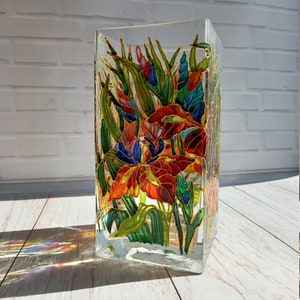 Op de afbeelding: Een heldere, vierkante glazen vaas met een handgeschilderd bloemenontwerp. De bloemen zijn voornamelijk rood, oranje en blauw, met groene bladeren en gouden contouren. De vaas staat op een lichtgekleurd houten oppervlak.