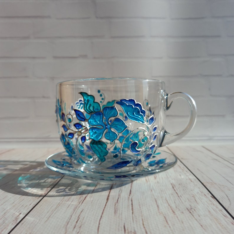 May include: A clear glass teacup and saucer set. The cup and saucer are decorated with a hand-painted floral design in shades of turquoise and blue. The floral design includes flowers, leaves, and vines. The cup has a handle.