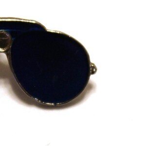 May include: A silver and blue enamel pin of aviator sunglasses with a gold pin back.