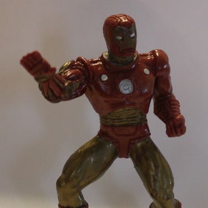 May include: A red and gold plastic action figure of Iron Man, a superhero from Marvel Comics. The figure is posed in a fighting stance with one arm raised and the other fist clenched. The figure has a red and gold suit with a silver chest plate and a red helmet with a gold faceplate.