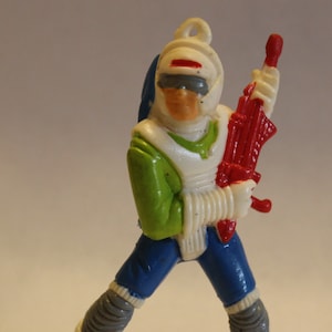 Puede incluir: Una figura de acción de plástico blanca, verde y azul de un explorador espacial que lleva un casco con una franja roja. La figura sostiene una pistola de plástico roja y blanca.