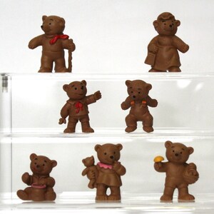 May include: Set of seven brown teddy bear figurines, each with a different outfit and accessory. The bears are arranged on a clear shelf.