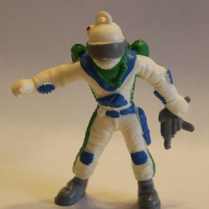 Puede incluir: Una figura de acción de astronauta blanca con un traje espacial verde y azul, sosteniendo una pistola gris. La figura tiene un casco gris con un punto rojo en la parte superior.