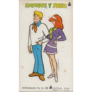 May include: A vintage sticker featuring cartoon characters Daphne and Fred from the Scooby-Doo franchise. The sticker is printed on a white background with the text "Daphne y Fred" in red and blue lettering. The characters are depicted in their signature outfits, with Daphne wearing a purple dress and Fred wearing a white shirt and blue pants.