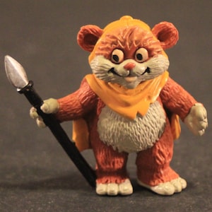 Puede incluir: Figura de acción Ewok vintage, con un cuerpo peludo de color marrón anaranjado, una capucha y bufanda amarillas y una lanza. La figura tiene una barriga blanca, patas blancas y una cara sonriente. Un juguete coleccionable.