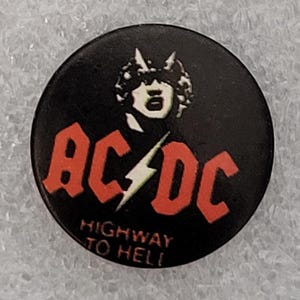 Puede incluir: Un botón redondo negro con el logo de la banda AC/DC en rojo y un rayo blanco. La imagen de una figura con cuernos está encima del logo. El texto "HIGHWAY TO HELL" está debajo del logo.