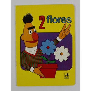 May include: A vintage children's book with a yellow cover featuring a cartoon character holding up two fingers. The book's title, "2 flores," is written in red and blue. The illustration includes two flowers in a red pot.