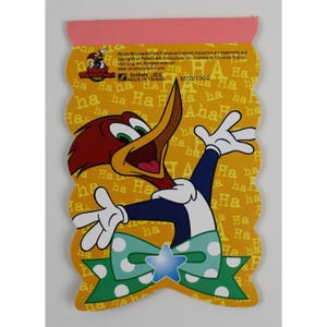 Op de afbeelding: Een geel notitieboekje met een cartoon afbeelding van Woody Woodpecker die een groene strik met witte stippen en een blauwe ster draagt. De achtergrond is geel met het woord "Ha" dat in verschillende groottes wordt herhaald. De tekst "Woody Woodpecker and Friends and related characters are trademarks and copyrights of Walter Lantz Productions, Inc. Licensed by Universal Studios Licensing, Inc. All rights reserved. www.universalstudios.com" staat bovenaan het notitieboekje gedrukt. De tekst "Shinn Jee Made in Taiwan" staat onder de afbeelding gedrukt.