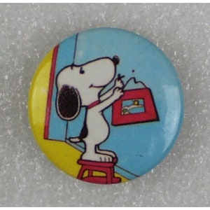Puede incluir: Un botón redondo con una imagen de dibujos animados de Snoopy pintando un cuadro en una pared. Snoopy está de pie en un taburete rojo y sostiene un pincel en su pata derecha. El fondo es azul y amarillo.