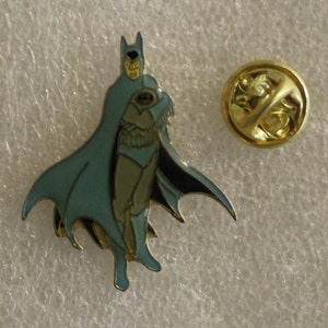 May include: A gold-colored enamel pin featuring a blue and black illustration of Batman in a cape. The pin has a safety clasp on the back.
