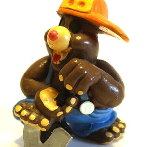 May include: A brown toy mole wearing a yellow hard hat with a blue jumpsuit and holding a silver shovel.