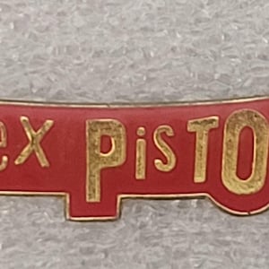 May include: A rectangular pin with the words "Sex Pistols" in gold lettering on a red background. The pin has a gold border and is likely a collectible item or a piece of memorabilia.