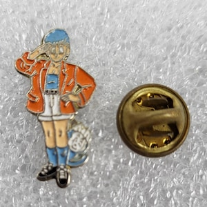 Puede incluir: Pin esmaltado con un personaje de dibujos animados con chaqueta naranja, gorro azul y pantalones cortos blancos, saludando. El pin tiene un respaldo dorado con un cierre de alfiler.