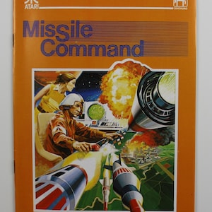 May include: A vintage Atari Missile Command game instruction manual with an orange cover and a colorful illustration of a futuristic scene with missiles, explosions, and a pilot in a cockpit.