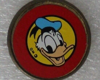 Peut inclure: Une épingle ronde en métal avec un fond rouge et un cercle jaune au centre. Le cercle présente une image de bande dessinée de Donald Duck avec un chapeau bleu, souriant et regardant vers la droite. Le texte "OWD" est visible dans le cercle jaune.
