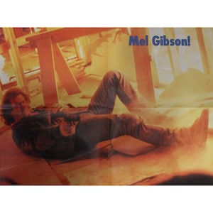 May include: A man lies on a wooden floor, holding a gun in his right hand. He is wearing blue jeans and a dark jacket. The background is a blurry image of a room with wooden beams and a bright orange glow. The text "Mel Gibson!" is visible in the upper right corner of the image.