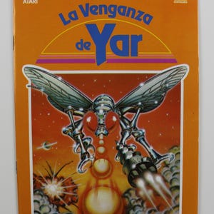 Könnte beinhalten: Ein Vintage-Atari-Videospiel-Kartuschencover mit orangefarbenem Hintergrund. Das Cover zeigt ein silbernes und rotes außerirdisches Insekt mit großen Augen und einer Waffe. Der Titel des Spiels ist "La Venganza de Yar" auf Spanisch.