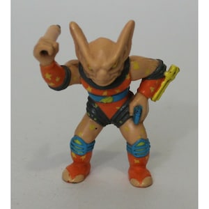 May include: A small, orange, blue and yellow plastic action figure of a creature with bat-like ears and a pointed nose. The figure is holding a yellow weapon in one hand and a long, thin, yellow weapon in the other.