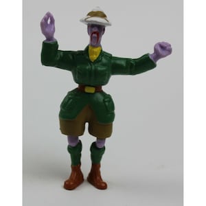 May include: A small, plastic figurine of a zombie dressed in a green safari outfit, complete with a khaki hat and shorts. The zombie has purple skin, a yellow collar, and brown boots. Its arms are outstretched.