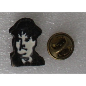 May include: Black and white enamel pin featuring a portrait of a person wearing a hat. The pin has a gold metal backing with a safety clasp.