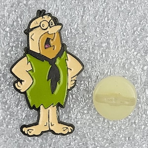 Puede incluir: Un colorido pin de esmalte con Pedro Picapiedra con los brazos en jarras, con una túnica verde y corbata negra. Junto a él, un pequeño pin redondo amarillo pálido con una ranura horizontal. Los pines están sobre un fondo plateado texturizado.