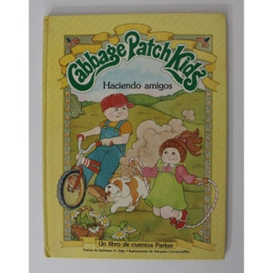 Könnte beinhalten: Ein gelber Buchumschlag mit dem Titel "Cabbage Patch Kids" in grüner und weißer Schrift. Das Buch zeigt eine Illustration von zwei Cabbage Patch Kids Puppen, eine fährt ein rotes Fahrrad und die andere springt Seil. Der Text "Haciendo amigos" ist in Spanisch unter dem Titel geschrieben. Das Buch trägt den Titel "Un libro de cuentos Parker" und enthält die Namen des Autors und des Illustrators.