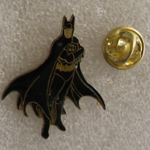 May include: A black and gold Batman pin with a black cape and a gold belt. The pin has a gold back with a safety clasp.