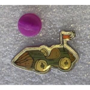Puede incluir: Un pin de metal plateado con una imagen de dibujos animados de un coche hecho de troncos de madera. El coche tiene una bandera roja en la parte trasera y ruedas amarillas. El pin está sobre un fondo blanco.