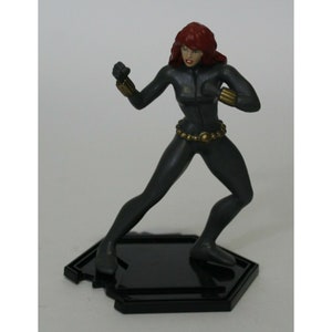 May include: Black and gray action figure of a female superhero with red hair. The figure is posed in a fighting stance with one leg forward and one arm raised. The figure is standing on a black hexagonal base.
