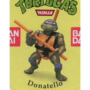 May include: Donatello, a Teenage Mutant Ninja Turtle, is depicted in a green and orange costume with a purple mask and a bo staff. The image is from a vintage trading card with the text "TURTLES NINJA" and "Donatello" on it.