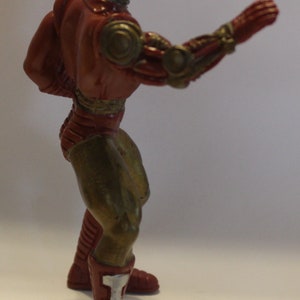 May include: A red and gold robot action figure with a metallic finish. The figure is posed with one arm raised and the other arm bent at the elbow. The figure is wearing red boots with white accents.