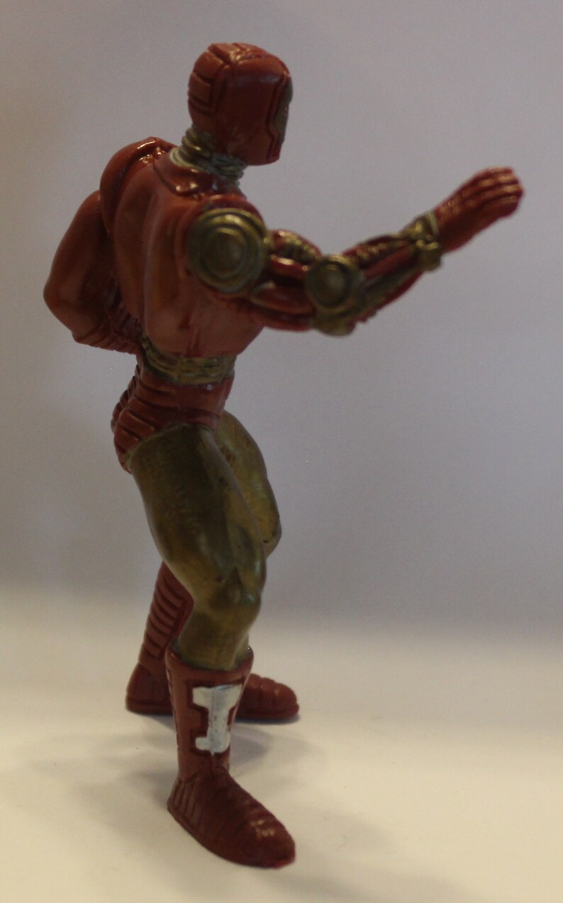 May include: A red and gold robot action figure with a metallic finish. The figure is posed with one arm raised and the other arm bent at the elbow. The figure is wearing red boots with white accents.